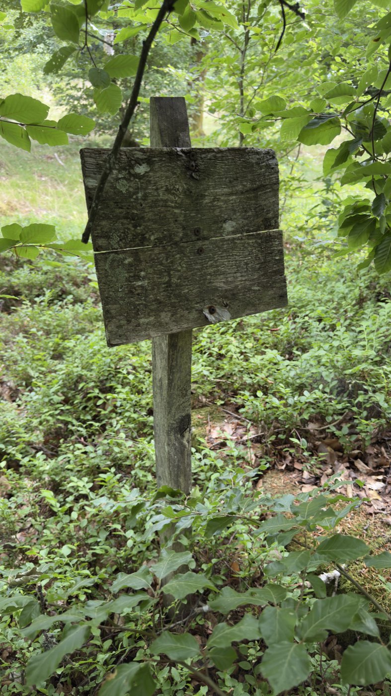Vollständig verwitterte, unlesbare Holztafel auf einem Pfosten neben dem Kreuz